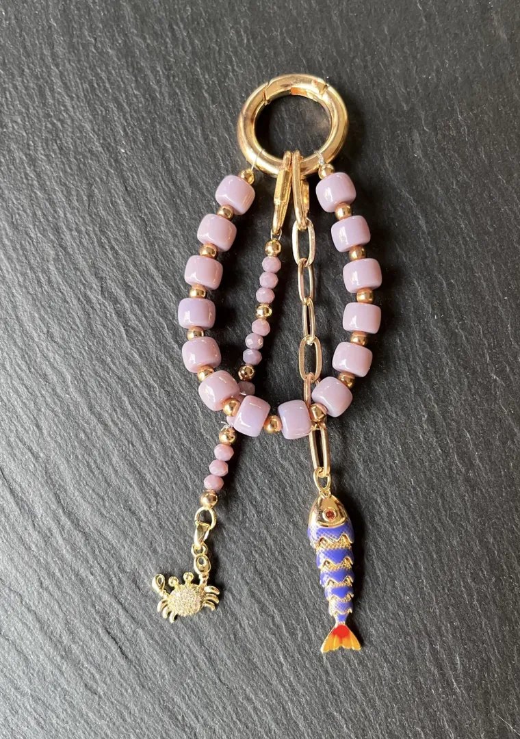 Unbearbeitetes Handyfoto eines handgemachten Schlüsselanhängers aus lila Perlen mit goldener Krabbe und blauem Fisch-Charm auf einer Schieferplatte